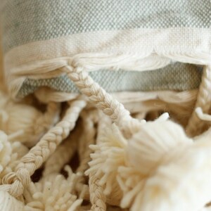 May include: A close-up of a light green throw blanket with white fringe and white pom-poms.