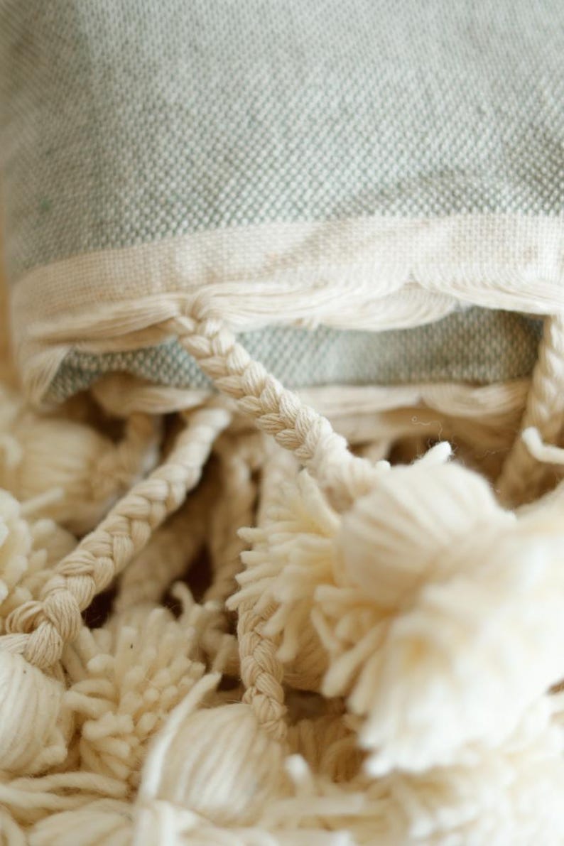 May include: A close-up of a light green throw blanket with white fringe and white pom-poms.