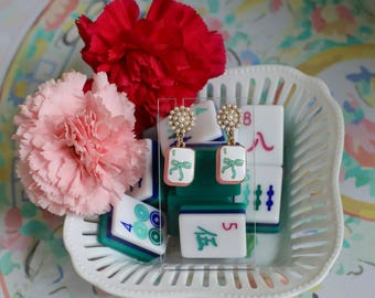May include: A pair of earrings with a pearl cluster top and a rectangular pendant featuring a green bow design. The earrings are displayed on a clear stand, surrounded by Mahjong tiles and pink and red carnations.