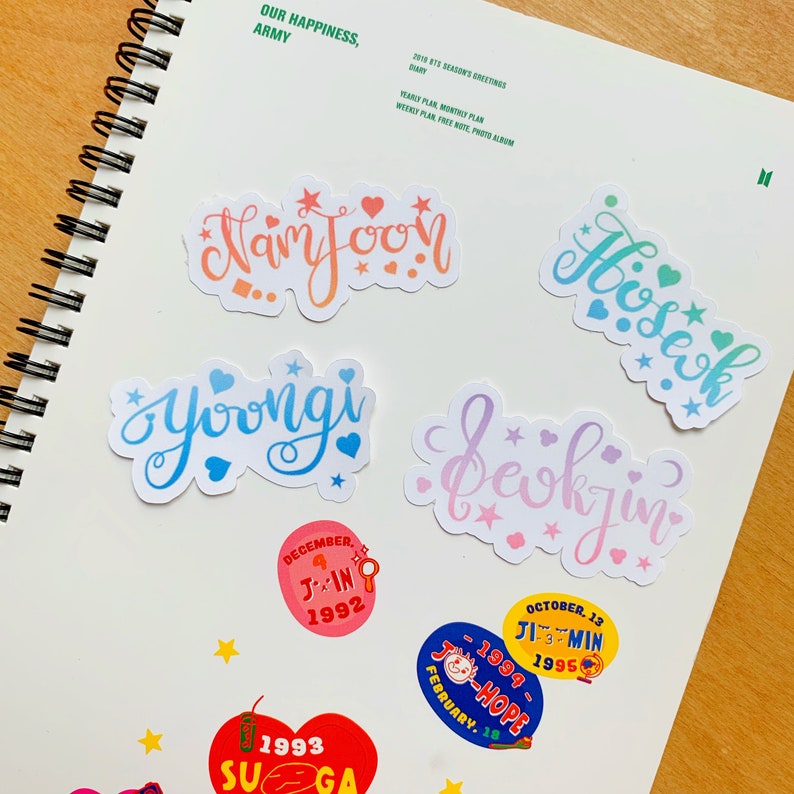 May include: A set of colorful stickers with the names of the members of the K-pop group BTS. The stickers are on a white background with a notebook page visible in the background. The stickers are in a variety of colors, including pink, blue, green, and orange. The names on the stickers are: Namjoon, Hoseok, Yoongi, Seokjin, Jin, Jimin, J-Hope, and Suga.