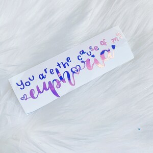 May include: A rectangular sticker with the words "You are the cause of my euphoria" in blue and iridescent pink lettering. The sticker has a white background and is decorated with small heart shapes.