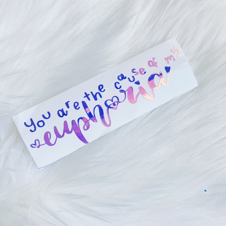 May include: A rectangular sticker with the words "You are the cause of my euphoria" in blue and iridescent pink lettering. The sticker has a white background and is decorated with small heart shapes.