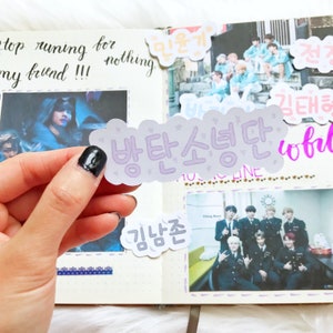 May include: A purple sticker with the Korean text "방탄소년단" (meaning "BTS") in a cloud-like shape. The sticker is held in a hand with black nail polish.