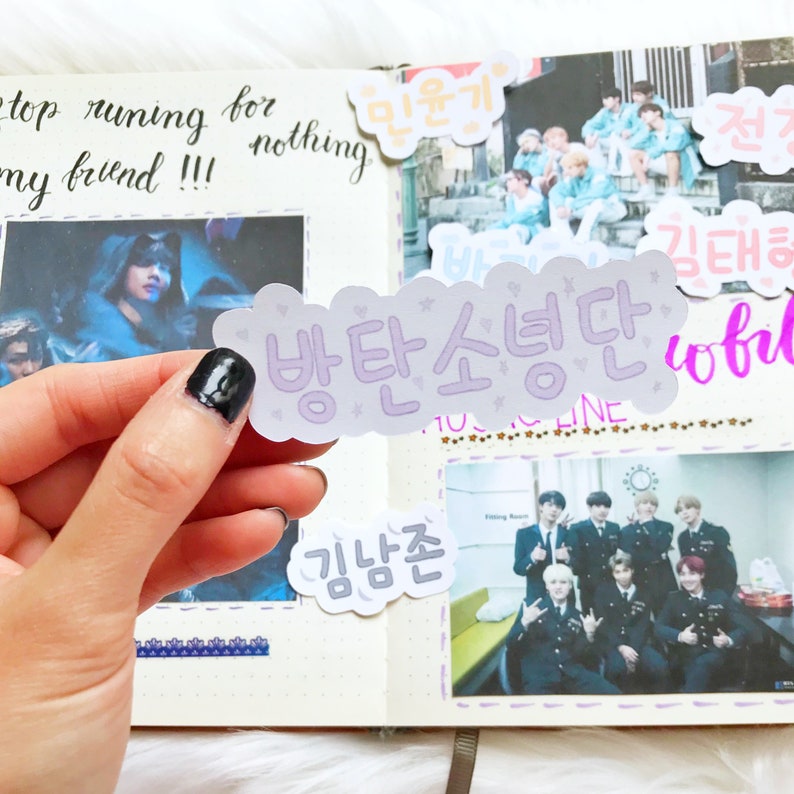 May include: A purple sticker with the Korean text "방탄소년단" (meaning "BTS") in a cloud-like shape. The sticker is held in a hand with black nail polish.