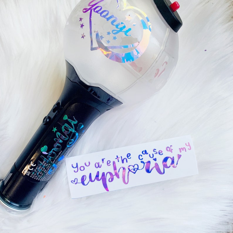 May include: A black and white light stick with the name "Yoongi" in holographic lettering. The light stick has a clear, spherical top with a red button and a heart-shaped design. A white card with the text "You are the cause of my euphoria" is also present.