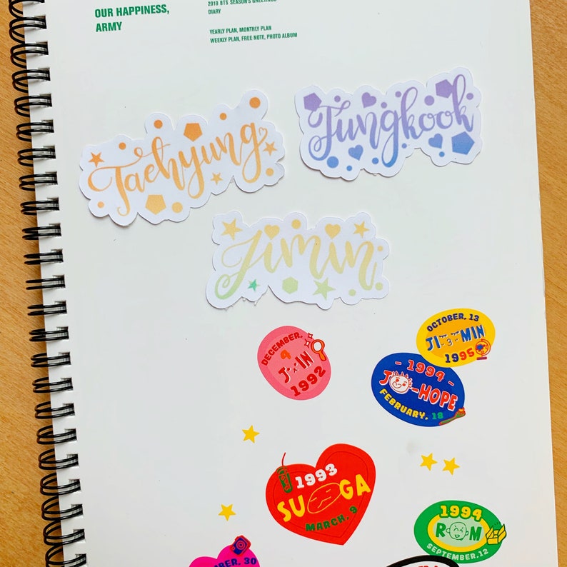 May include: A collection of colorful stickers featuring the names of the members of the South Korean boy band BTS. The stickers are in a variety of shapes and sizes, and each one features a different member's name in a unique font. The stickers are perfect for adding a touch of personality to laptops, water bottles, or journals.