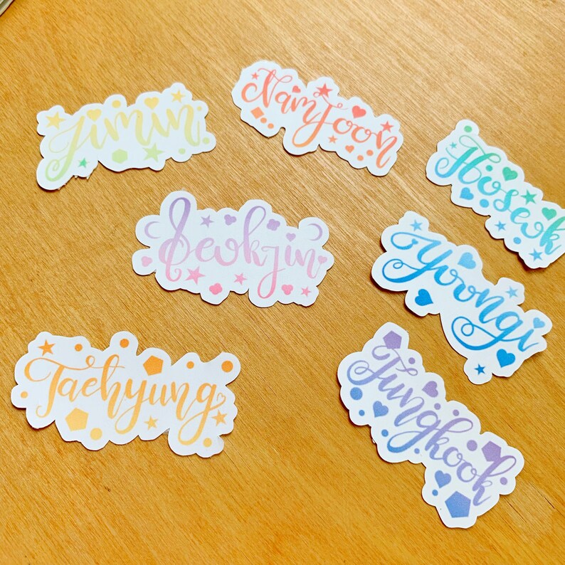 May include: Seven colorful stickers with the names of the members of the K-pop group BTS: Jimin, Namjoon, Hoseok, Seokjin, Yoongi, Taehyung, and Jungkook. Each sticker has a different color and features a unique design with stars and hearts.