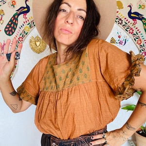 Peut inclure: Haut orange-brun à manches courtes et bordure à volants. Le haut a une encolure carrée avec un motif floral vert. Le modèle porte un short en jean marron foncé et un chapeau beige. Une tapisserie colorée est en arrière-plan.