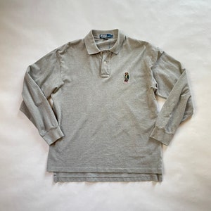 Puede incluir: Un polo de manga larga gris claro con cuello y tres botones. La camisa presenta un pequeño oso bordado con una chaqueta. El nombre de la marca es visible en el cuello.