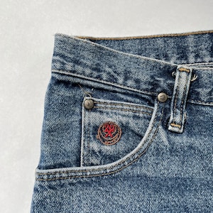 May include: Close-up of a blue denim pocket with a small red embroidered logo. The logo features a circle with a cross inside and the number 20 above it.