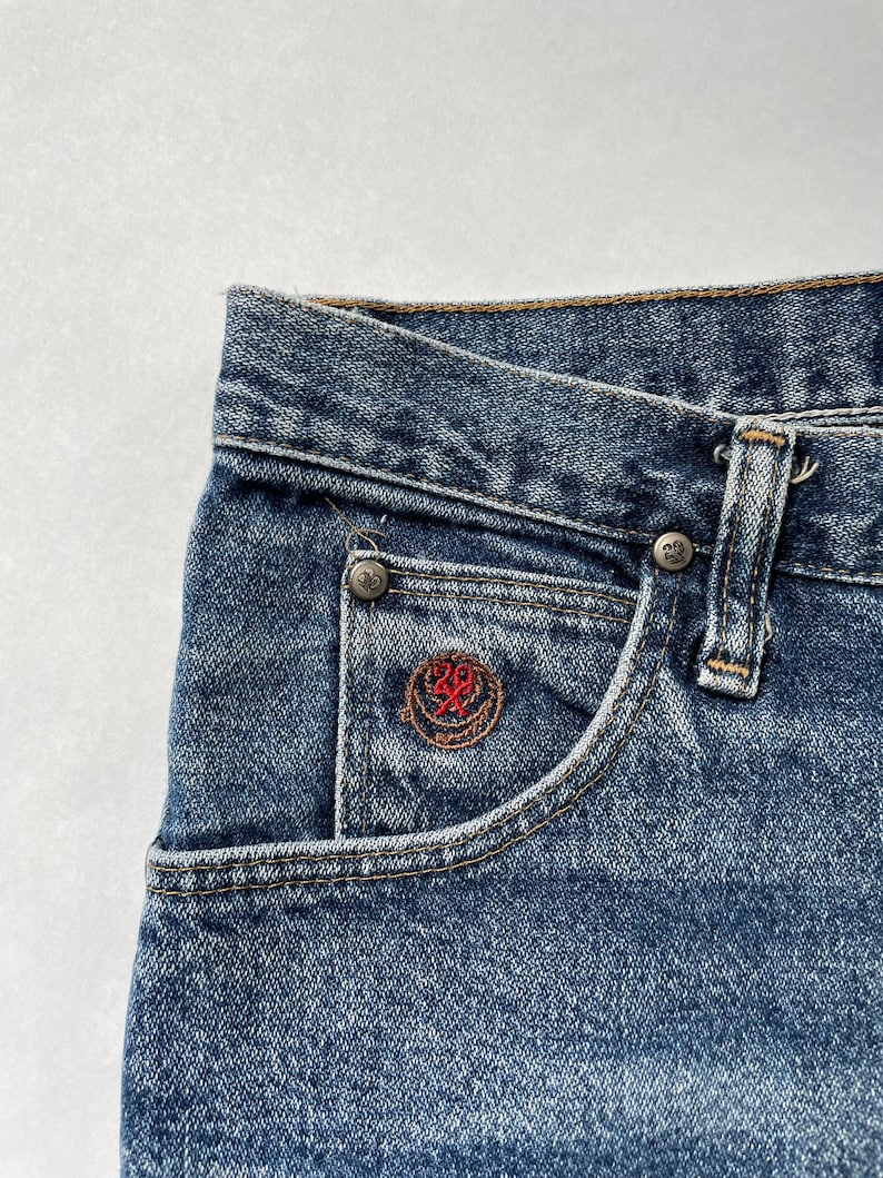 May include: Close-up of a blue denim pocket with a small red embroidered logo. The logo features a circle with a cross inside and the number 20 above it.