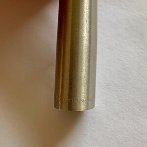 May include: Close-up of a metallic pen with a brushed finish. The text "MADE IN SPAIN" is engraved near the base of the pen. The pen has a cylindrical shape and a silver color.