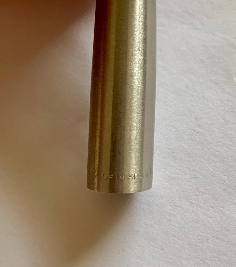 May include: Close-up of a metallic pen with a brushed finish. The text "MADE IN SPAIN" is engraved near the base of the pen. The pen has a cylindrical shape and a silver color.