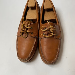 May include: A pair of brown leather boat shoes with laces. The shoes have a white stitching detail around the edges.