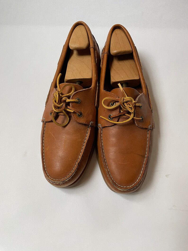 May include: A pair of brown leather boat shoes with laces. The shoes have a white stitching detail around the edges.