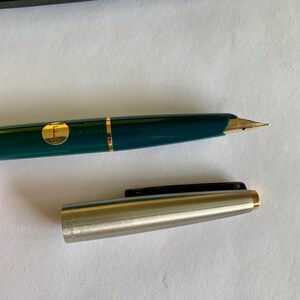 May include: A teal fountain pen with gold accents and a gold nib. The pen has a gold band and a circular gold emblem with the letter "F". The pen's cap is silver with a black clip and gold trim.