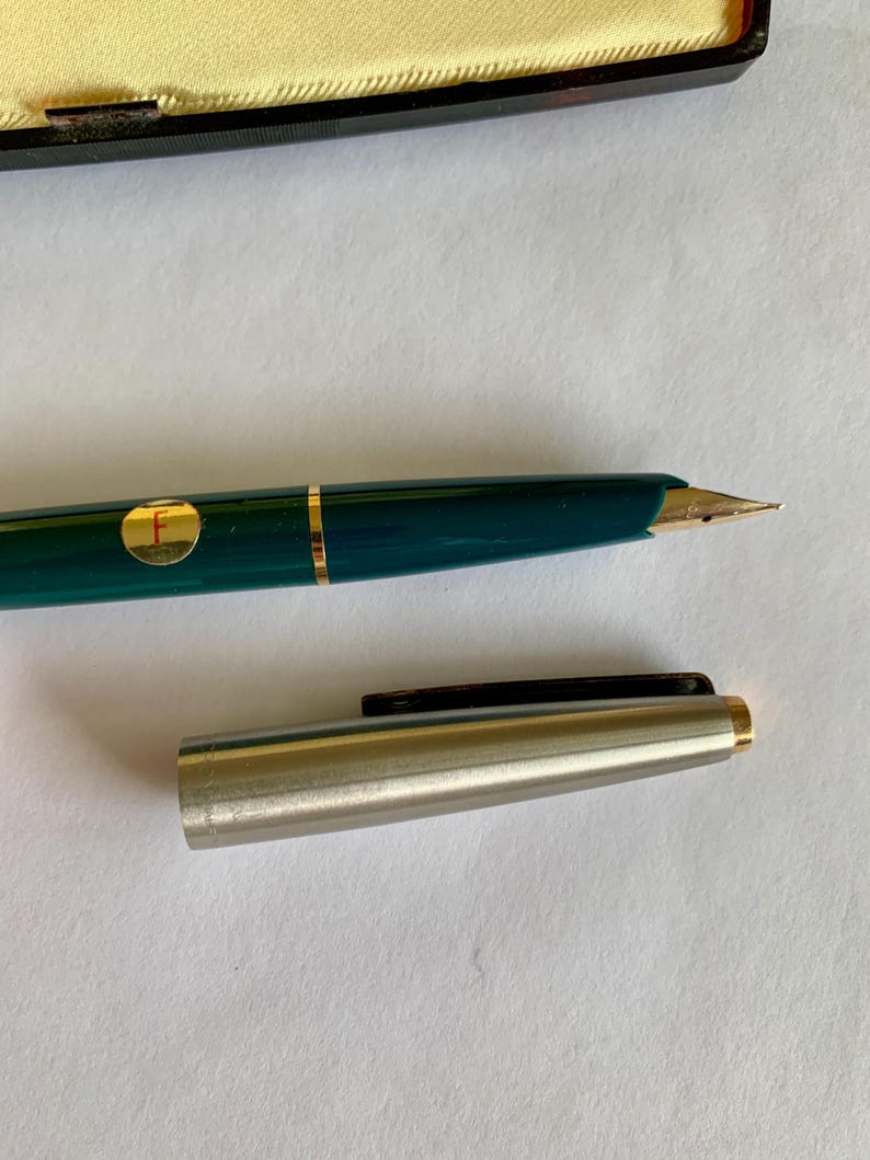 May include: A teal fountain pen with gold accents and a gold nib. The pen has a gold band and a circular gold emblem with the letter "F". The pen's cap is silver with a black clip and gold trim.