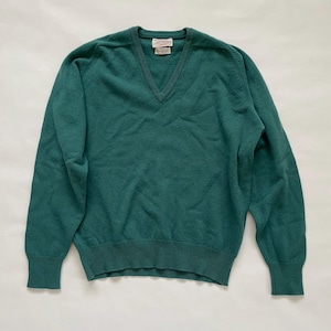 May include: A teal green V-neck sweater. The sweater has long sleeves and ribbed cuffs and hem. A small tag is visible at the back of the neck. The sweater is made of a soft, knitted material.