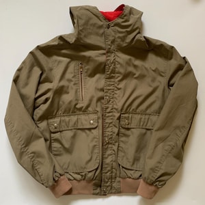 May include: A tan jacket with a red interior lining. The jacket has a high collar, a zipper pocket, and two flap pockets with button closures. The cuffs and waistband are a darker tan color.
