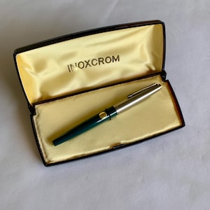 May include: A vintage INOXCROM fountain pen in its original box. The pen has a silver cap and a dark green barrel. The box is black with a gold satin interior and the brand name INOXCROM is printed in gold.
