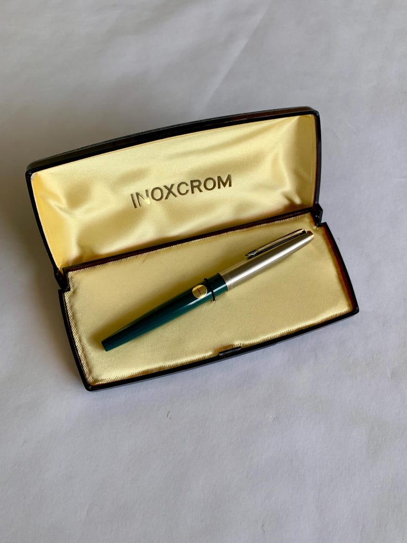 May include: A vintage INOXCROM fountain pen in its original box. The pen has a silver cap and a dark green barrel. The box is black with a gold satin interior and the brand name INOXCROM is printed in gold.
