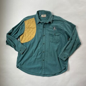 May include: A green long-sleeved button-down shirt with a yellow quilted patch on the left shoulder. The shirt has a small embroidered bird design on the right chest pocket.