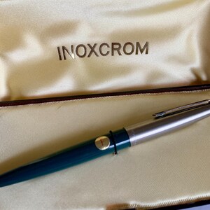 May include: A teal and silver INOXCROM fountain pen rests inside a cream-colored satin-lined case. The pen has a silver tip and a gold-colored band with the letter "F". The case is black with the brand name INOXCROM printed in gold.