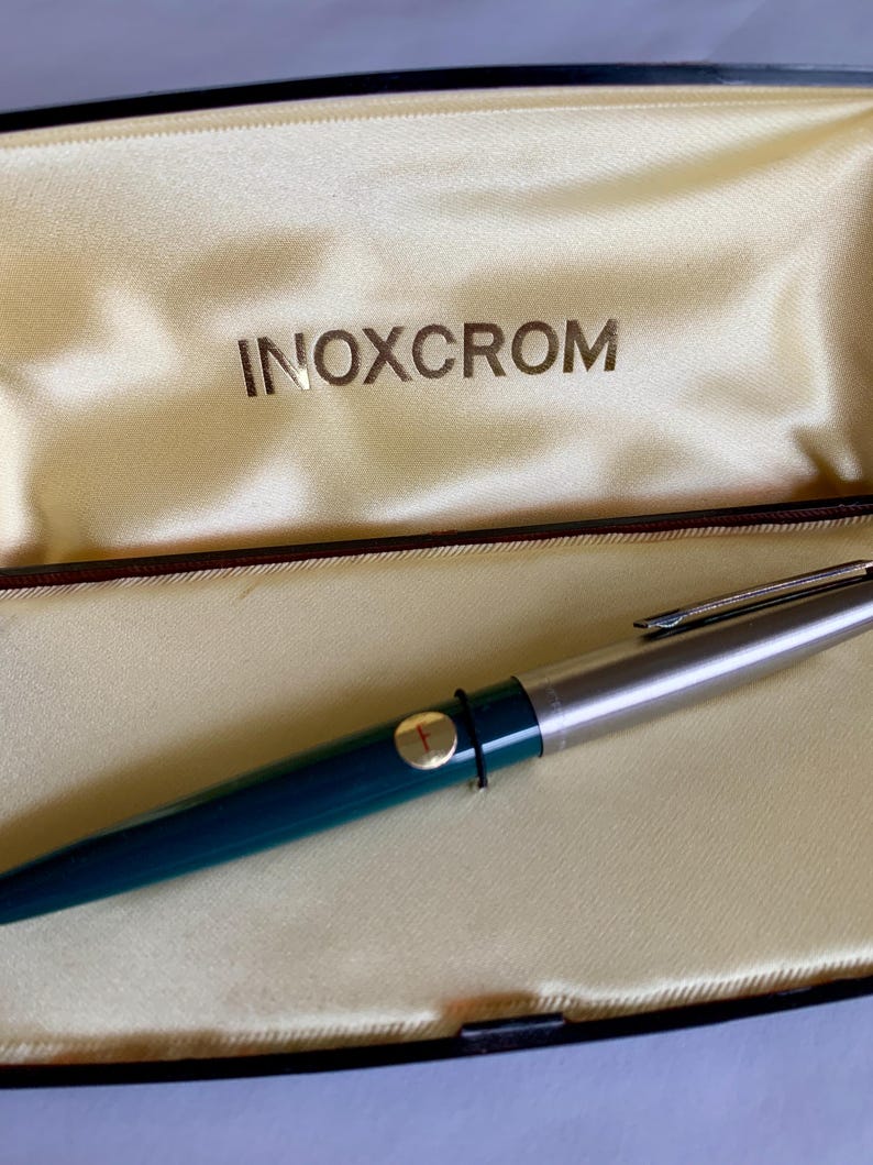 May include: A teal and silver INOXCROM fountain pen rests inside a cream-colored satin-lined case. The pen has a silver tip and a gold-colored band with the letter "F". The case is black with the brand name INOXCROM printed in gold.
