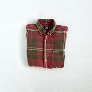 May include: A red and green plaid button-down shirt with a collar and a chest pocket. The shirt has the "GAP" logo on the collar. The shirt is folded neatly on a white surface.