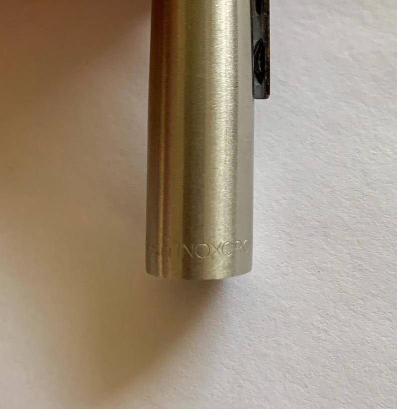 May include: Close-up of a silver-colored pen with the text "INOXCR" engraved on the barrel. The pen has a sleek, metallic finish and a black clip. The pen is likely a writing instrument.
