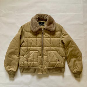 May include: A tan quilted jacket with a faux fur collar. The jacket has a zipper closure, two front pockets, and ribbed cuffs and hem. The label on the collar reads "Golden Fleece".