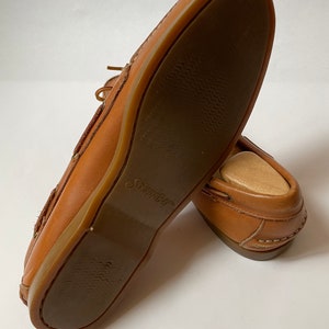 May include: A pair of brown leather boat shoes with a brown rubber sole. The sole has a textured pattern and the word "Swims" is printed on the sole.