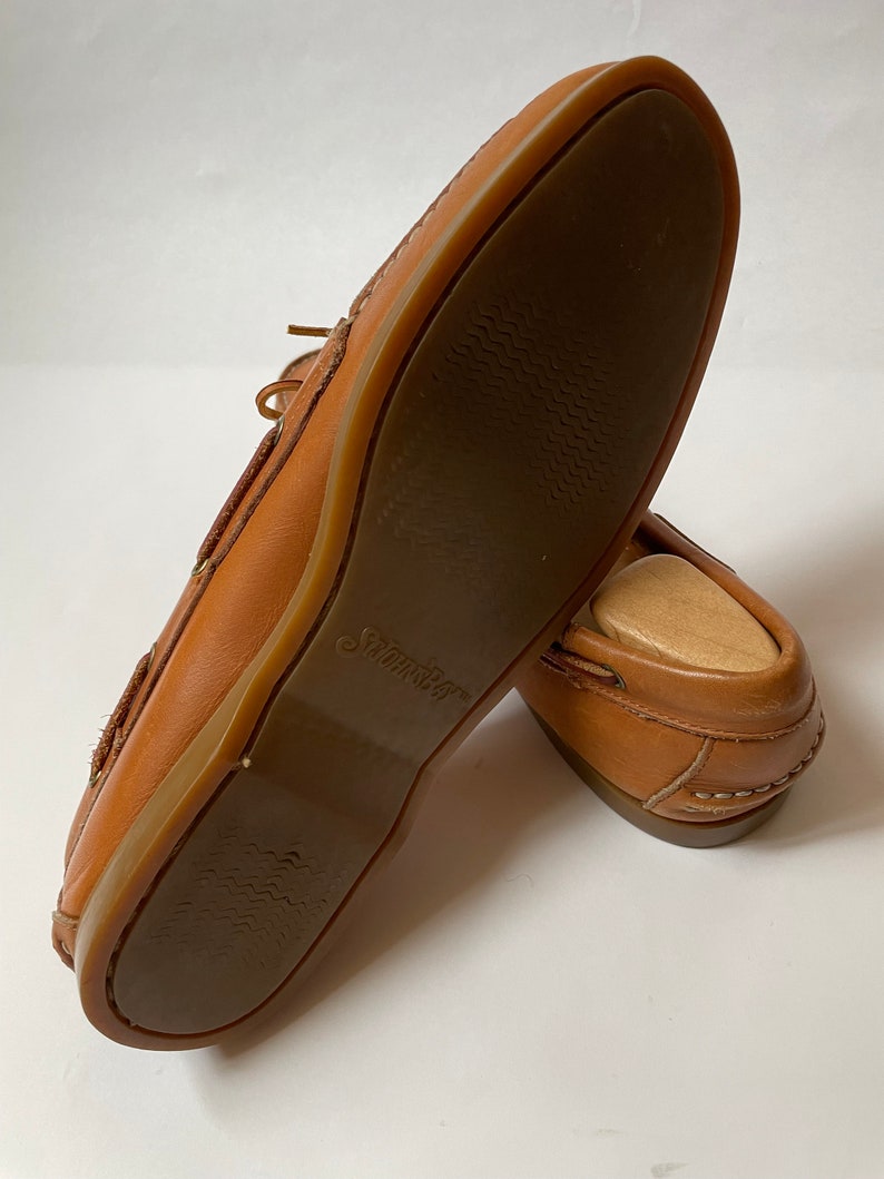 May include: A pair of brown leather boat shoes with a brown rubber sole. The sole has a textured pattern and the word "Swims" is printed on the sole.