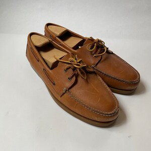 May include: A pair of brown leather boat shoes with laces. The shoes have a light brown sole and are worn.