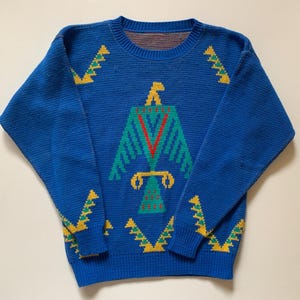 May include: A blue knit sweater with a crew neck and long sleeves. The sweater features a geometric design of a bird in shades of green, red, and yellow. Additional yellow geometric patterns are on the sleeves.