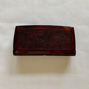 May include: A rectangular, dark brown box with rounded edges. The top surface has a textured, mottled appearance. The box appears to be made of a glossy material, possibly wood or a composite. The box is set against a plain, off-white background.