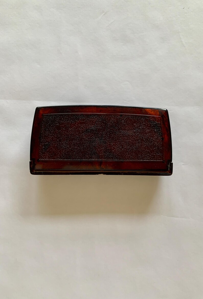 May include: A rectangular, dark brown box with rounded edges. The top surface has a textured, mottled appearance. The box appears to be made of a glossy material, possibly wood or a composite. The box is set against a plain, off-white background.