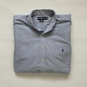 May include: A light blue button-down shirt with a pocket on the left chest. The shirt has a button-down collar and a small navy blue logo on the pocket. The shirt is folded neatly.