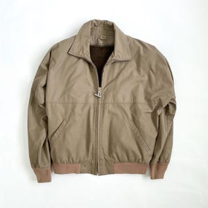 Op de afbeelding: Een bruine bomberjack met een volledige ritssluiting, twee voorzakken en een bruine geribde zoom en manchetten. De jas heeft een kraag en een zachte binnenvoering.