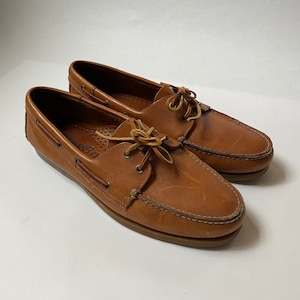 May include: A pair of brown leather boat shoes with laces. The shoes have a light brown sole and are made of a soft, supple leather.
