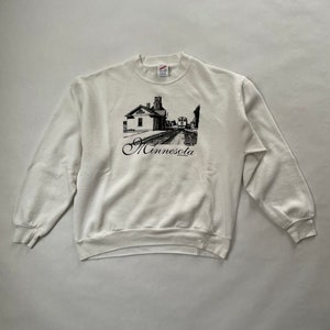 May include: White long-sleeve sweatshirt with a black and white graphic of a building and train tracks. The text "Minnesota" is printed in black cursive below the graphic.