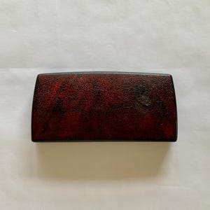 May include: A rectangular, dark red box with a textured surface. The box has rounded edges and a black border. The overall appearance suggests a vintage or antique item, possibly a case or container.