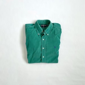 Puede incluir: Camisa verde azulado abotonada, doblada. Tiene cuello abotonado, botones blancos y un pequeño logo azul en el pecho. La tela parece pana o similar, con un ligero brillo. La camisa está doblada sobre una superficie blanca.