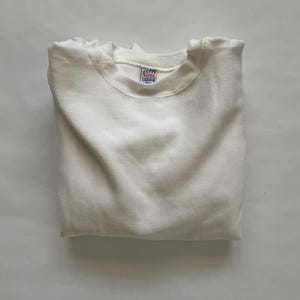 May include: A folded, off-white sweatshirt. The crew neck sweatshirt has a small tag with red and blue text. The garment is neatly folded, showcasing its clean lines and simple design. The fabric appears soft and comfortable.