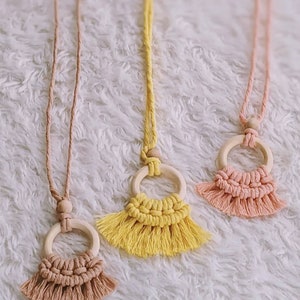 Puede incluir: Tres collares de macramé con anillos de madera y flecos. Los collares son de color rosa, amarillo y marrón.