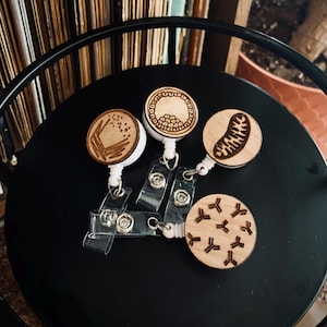 Può includere: Tre rotoli per badge in legno con diversi design. I design sono una casa, una cellula e un batterio. I rotoli sono su una superficie nera.