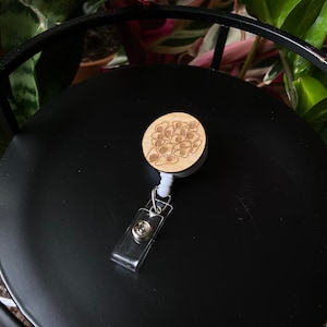 Może przedstawiać: Drewniany zwijany uchwyt na identyfikator z białym, chowanym sznurkiem i przezroczystym plastikowym uchwytem na identyfikator. Drewniana część ma laserowo grawerowany wzór skupiska oczu.