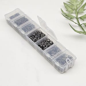Op de afbeelding: Een doorzichtige plastic organizer gevuld met verschillende maten zilverkleurige sprongringen. De organizer heeft meerdere compartimenten, elk met een andere hoeveelheid metalen ringen. De sprongringen worden gebruikt voor het maken van sieraden en andere ambachten.