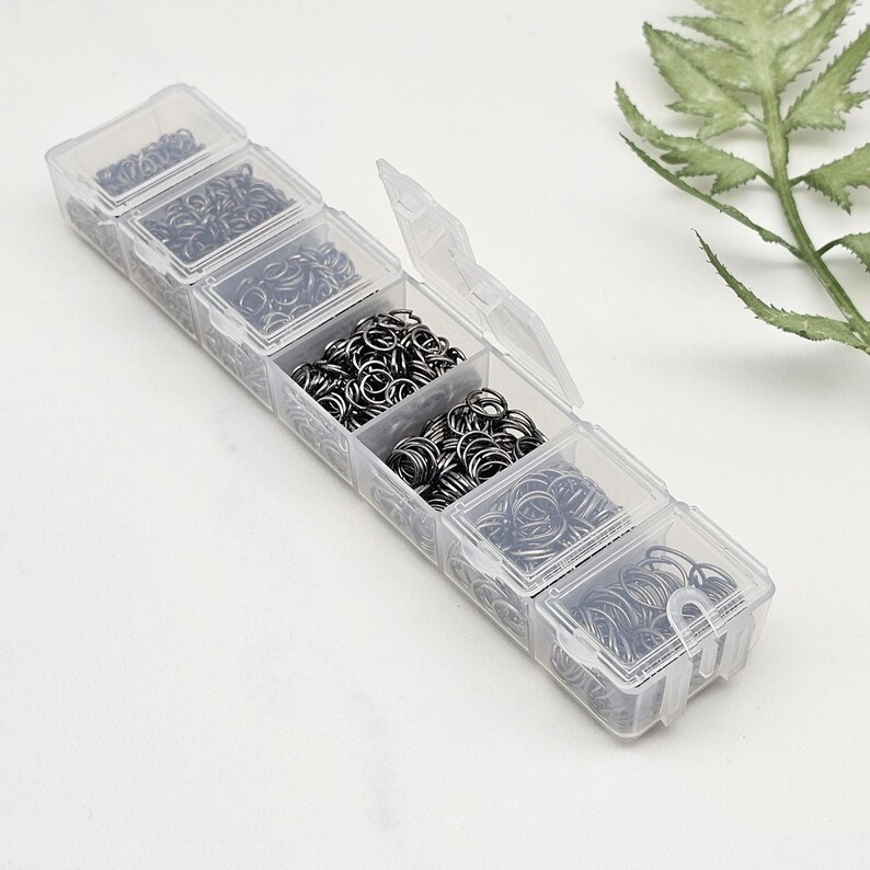Op de afbeelding: Een doorzichtige plastic organizer gevuld met verschillende maten zilverkleurige sprongringen. De organizer heeft meerdere compartimenten, elk met een andere hoeveelheid metalen ringen. De sprongringen worden gebruikt voor het maken van sieraden en andere ambachten.