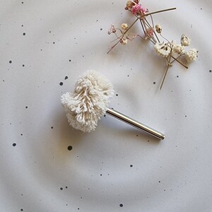 May include: A small, fluffy, cream-coloured object with a silver metal handle, possibly a jewellery cleaner or makeup tool, rests on a speckled, light grey surface. Delicate dried flowers with pink and white accents are also present.